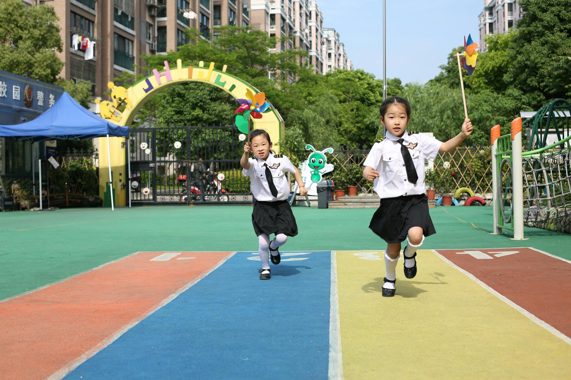 Dwoje dzieci biegnie po kolorowej bieżni z wiatraczkami na szkolnym podwórku